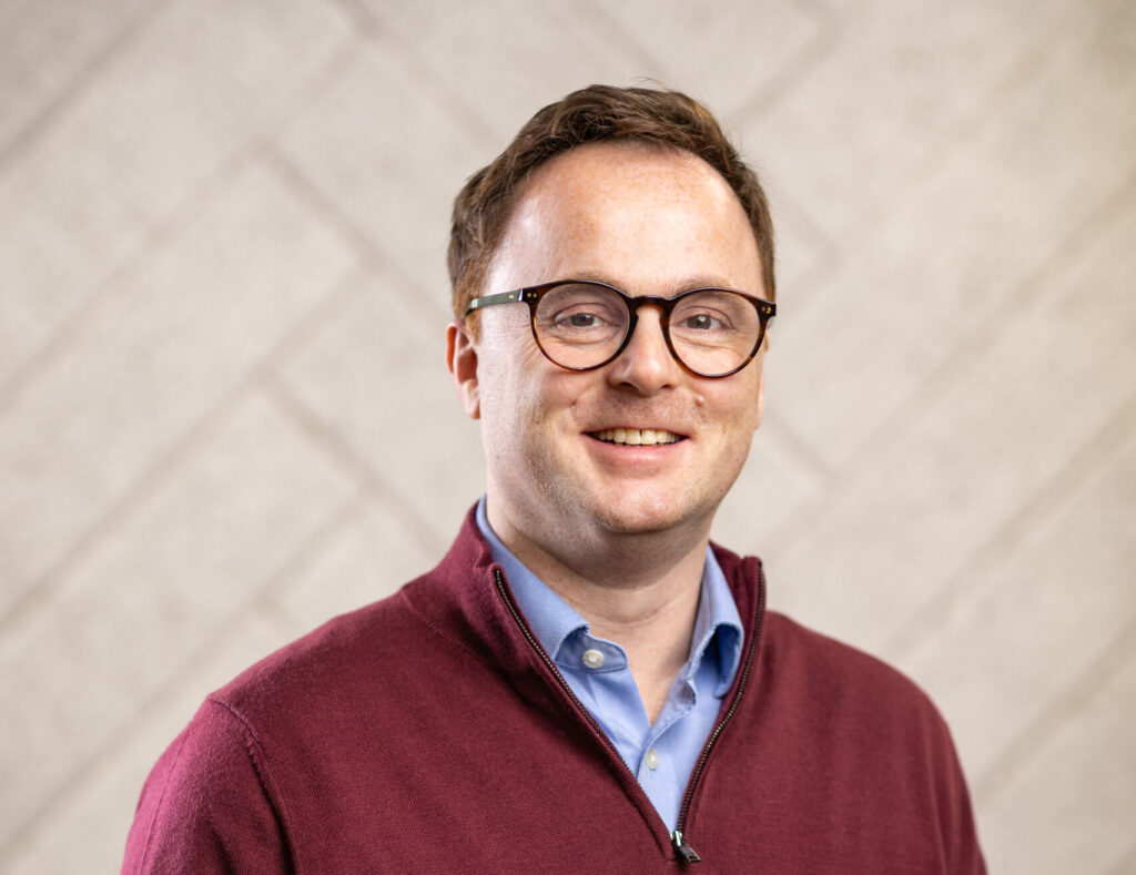 Man wearing glasses and a maroon zip-up sweater over a blue shirt smiling