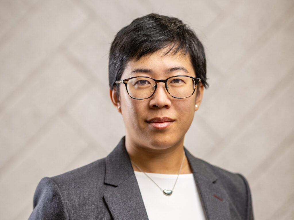 Professional headshot of a person with short black hair, glasses, and a gray blazer