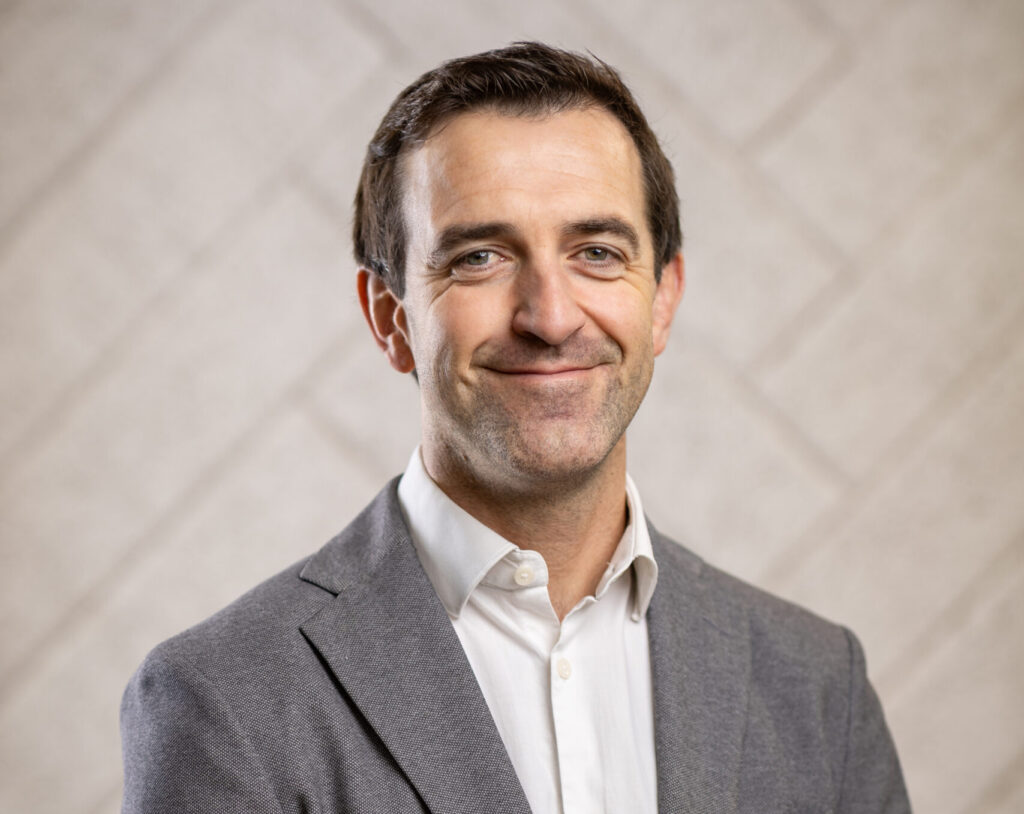 Man in gray blazer and white shirt smiling against a beige background